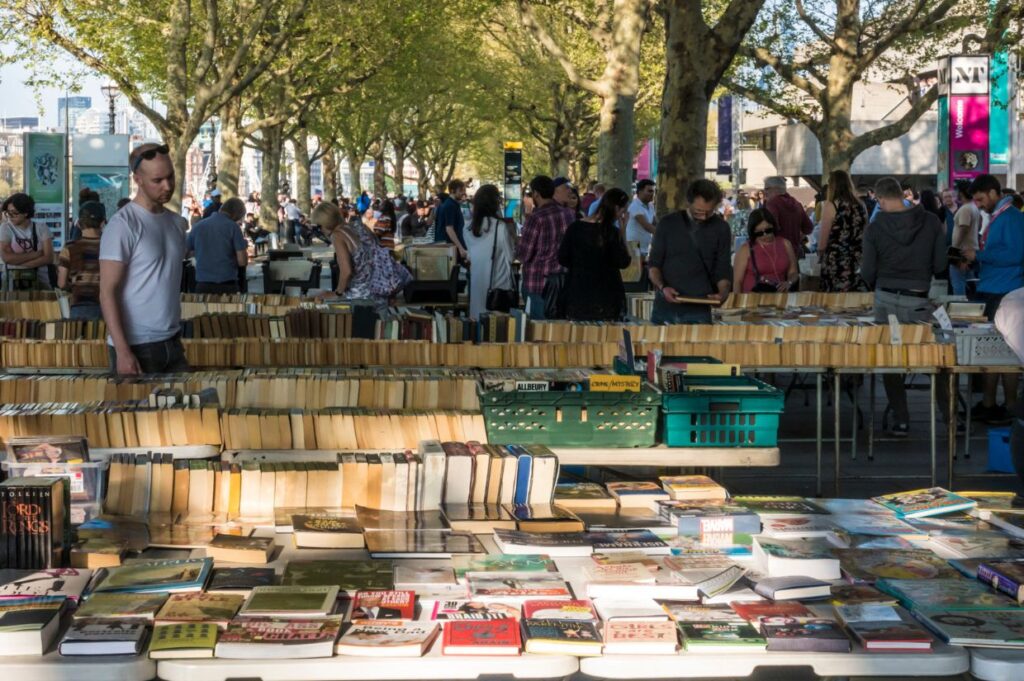 An outdoor book market on London’s South Bank under the shade of tall trees. Rows of tables filled with second-hand books stretch along the walkway, with people browsing through them.