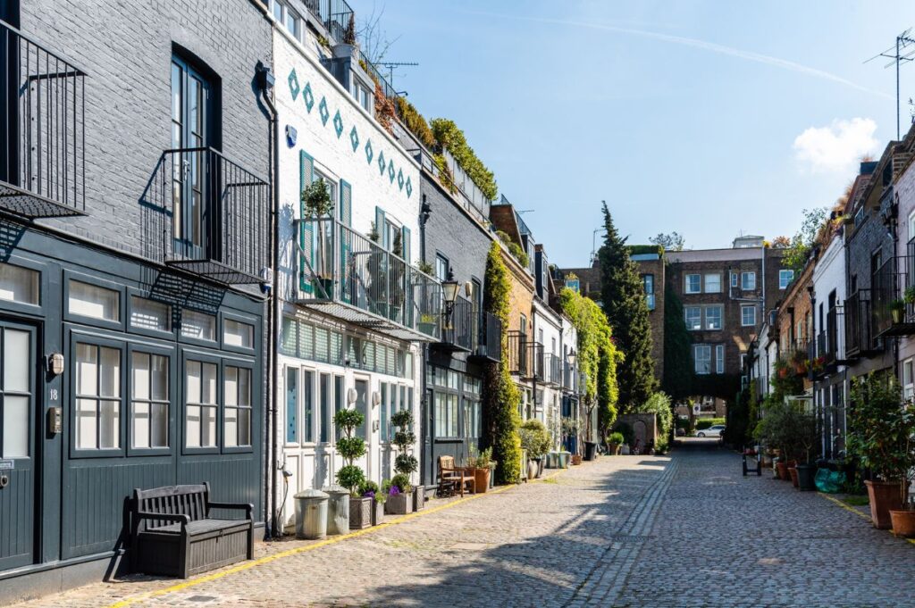 A charming cobblestone alley in St. Luke’s Mews, London, lined with colorful, ivy-clad houses featuring balconies, large windows, and potted plants. The peaceful street is bathed in sunlight, creating a picturesque and inviting atmosphere.