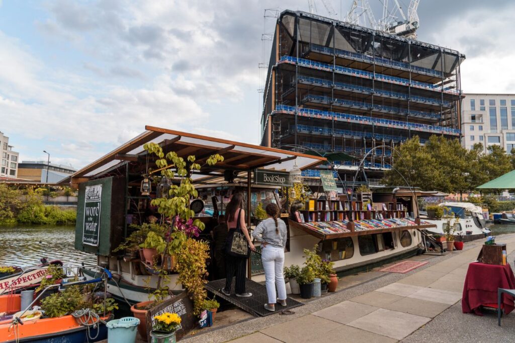 A floating bookshop on a canal with two visitors browsing books, set against a backdrop of a construction site and greenery.