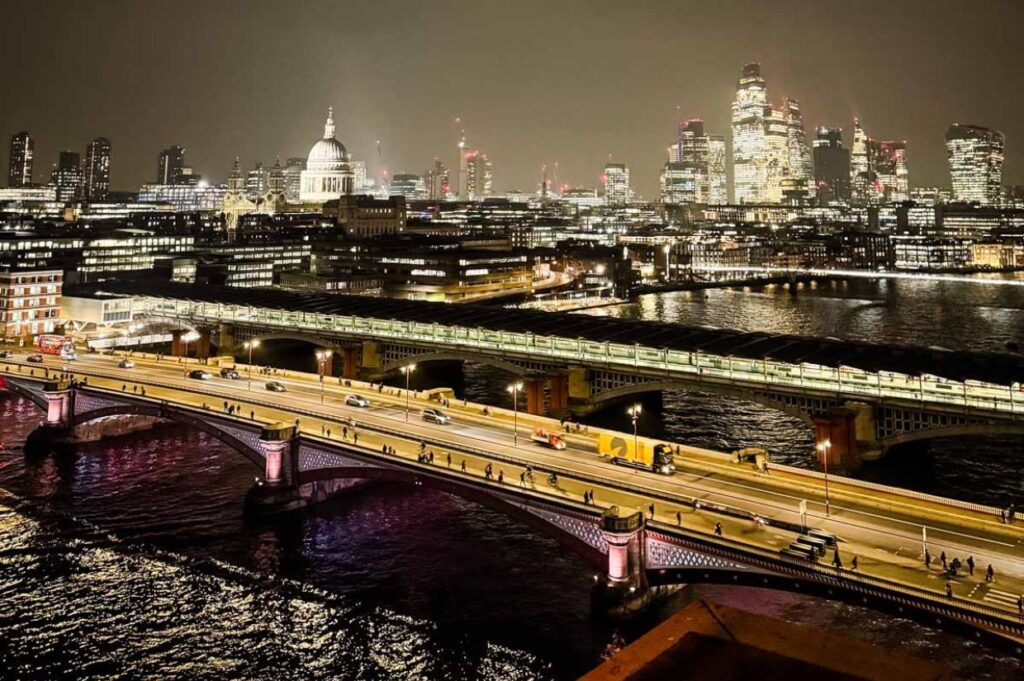 A nighttime cityscape, likely of London, featuring illuminated buildings and bridges spanning a river.
