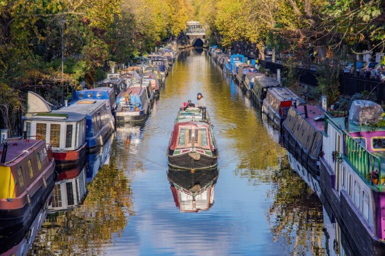 Colorful narrowboats line both sides of a peaceful canal in Little Venice, London, with trees reflecting on the calm water and a boat carrying two people moving through the center.