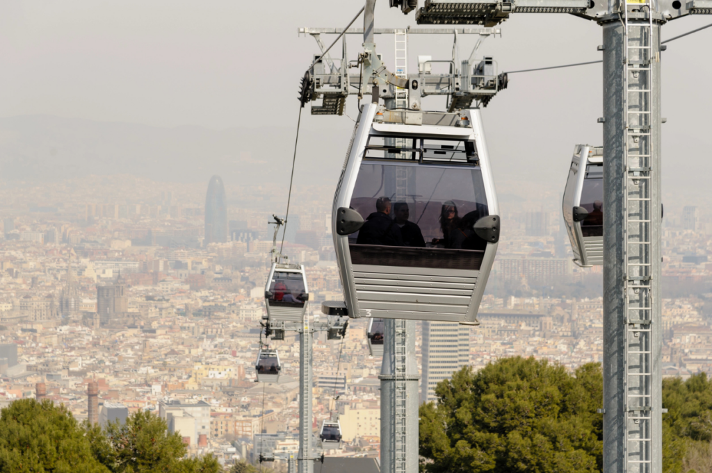 Cable cars glide over a cityscape, with modern buildings in the haze. Trees below and people inside convey a leisurely, panoramic experience.