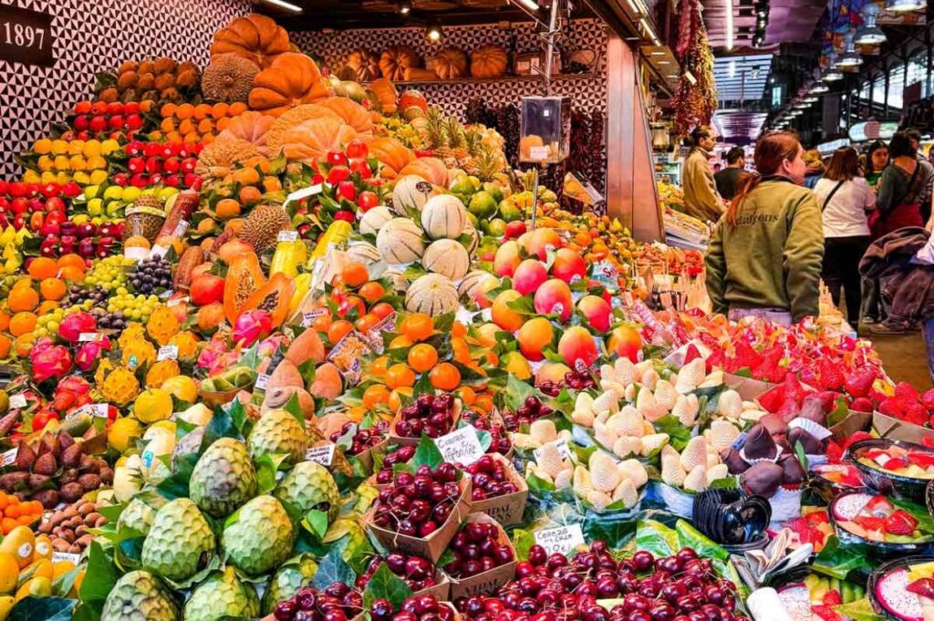 A vibrant market stall displays a colorful array of fruits, including oranges, melons, cherries, and papayas. People browse the lively, bustling market.