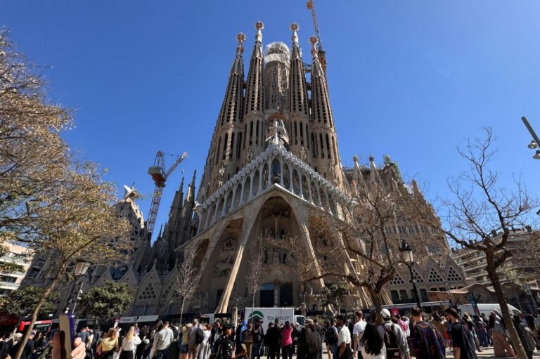 The image shows the Sagrada Familia in Barcelona under a clear blue sky. Crowds gather at its base, surrounded by cranes and leafless trees.