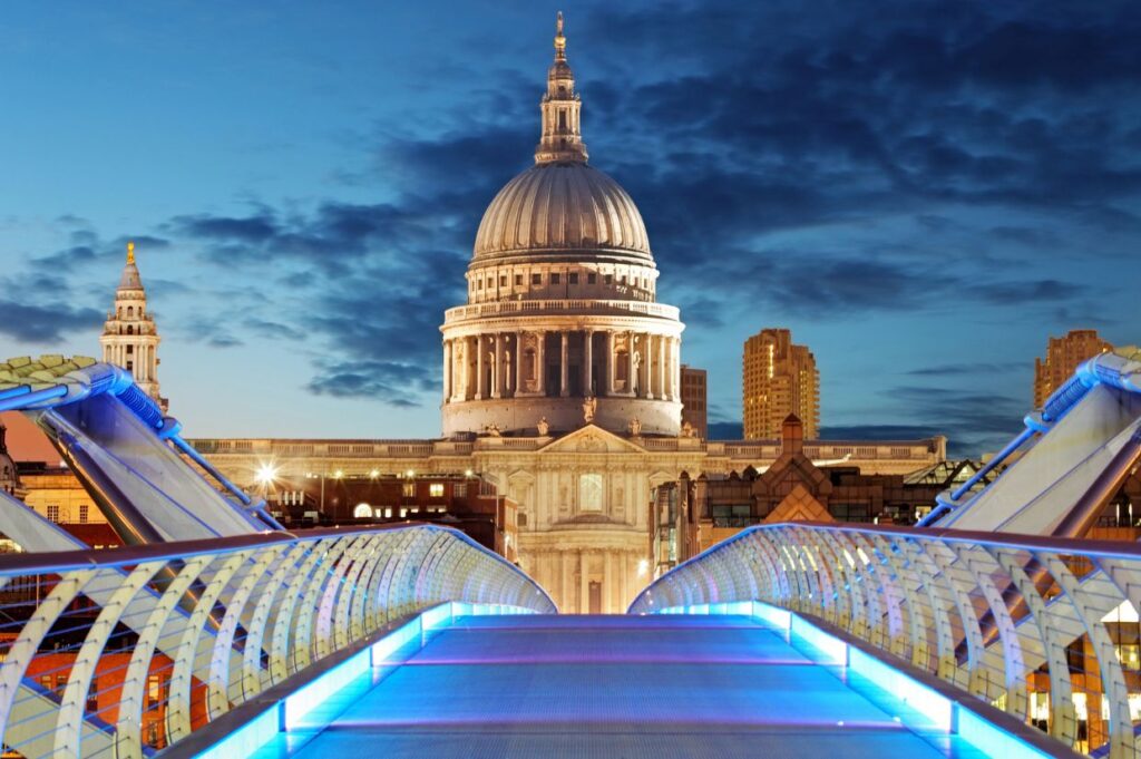 View of the Millennium Bridge in London at dusk, leading towards the illuminated dome of St. Paul's Cathedral under a dramatic evening sky.
