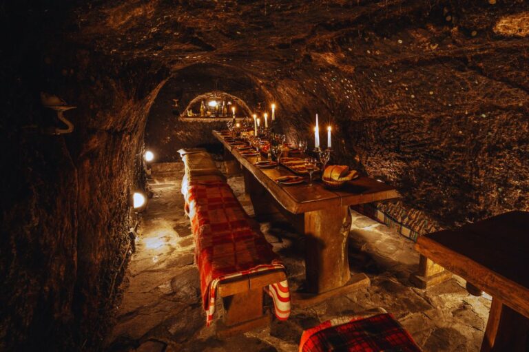 Rustic underground dining room with a wooden table set for a feast, lit by candles. Stone walls and benches with red plaid blankets create a cozy ambiance.