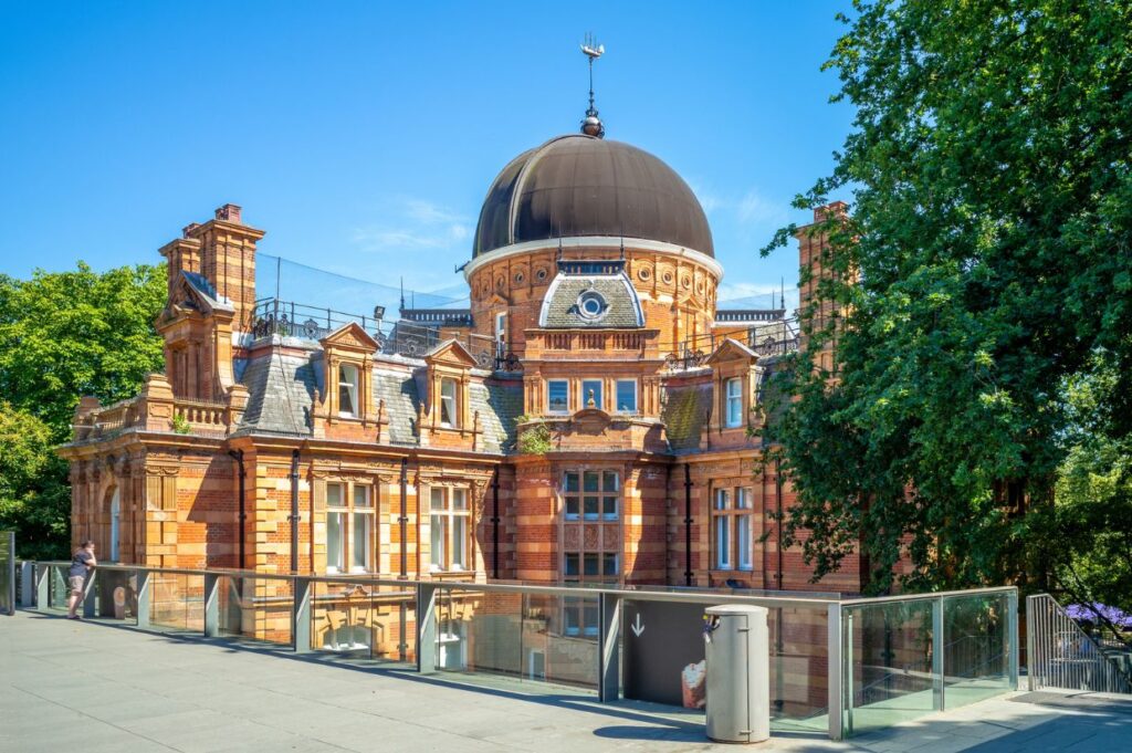 The historic Royal Observatory in Greenwich, London, featuring red-brick architecture with decorative stonework and a large black domed roof under a clear blue sky.
