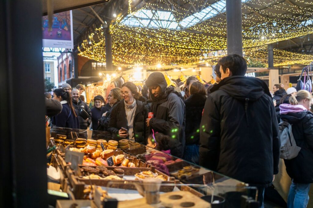 Busy scene at Spitalfields Market in London, with people browsing food stalls under a canopy of twinkling fairy lights.