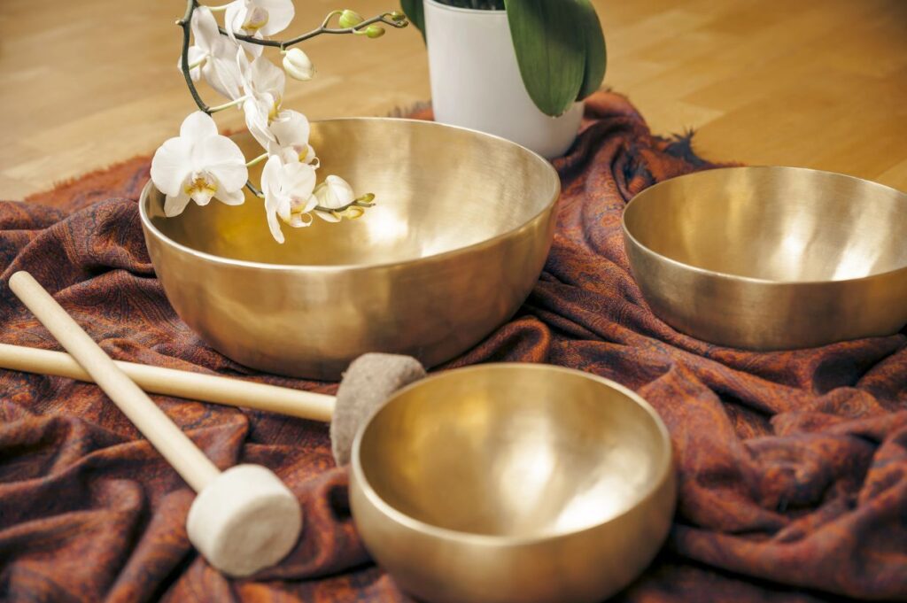 Brass singing bowls of varying sizes arranged on a patterned cloth, accompanied by wooden mallets and a white orchid branch, creating a calm and serene setting.