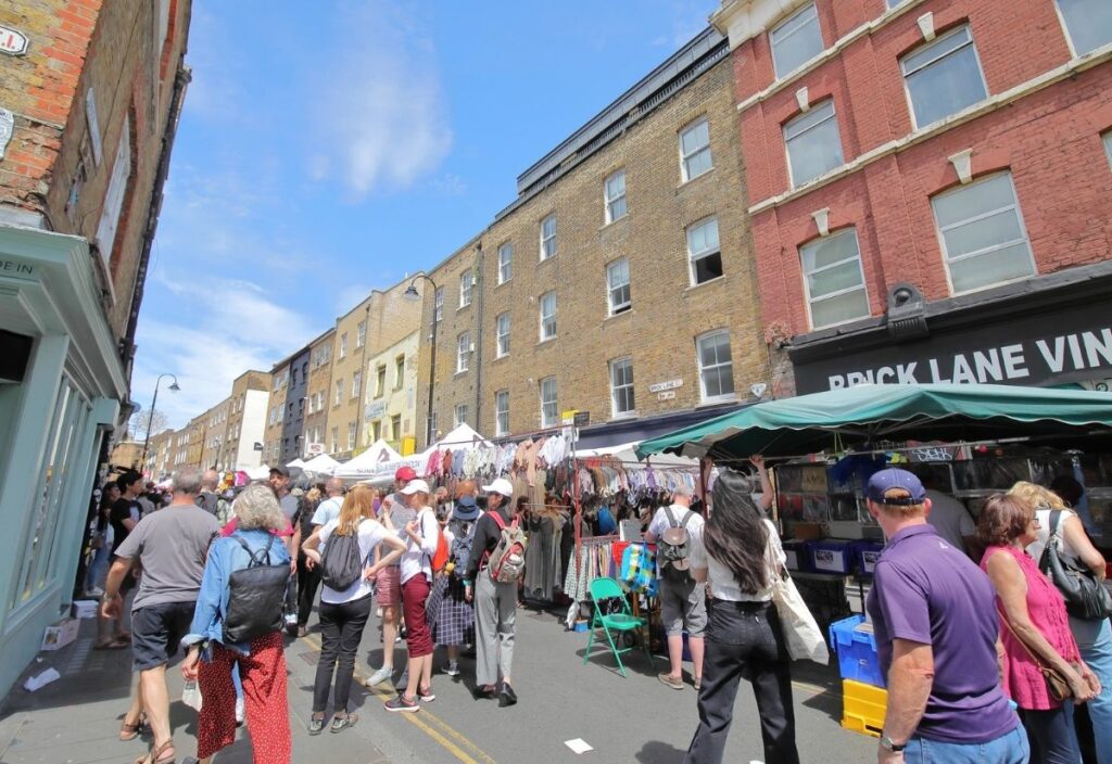 Bustling outdoor market on a sunny day at Brick Lane, London. Crowds browse stalls with colorful clothing, against a backdrop of historic brick buildings.