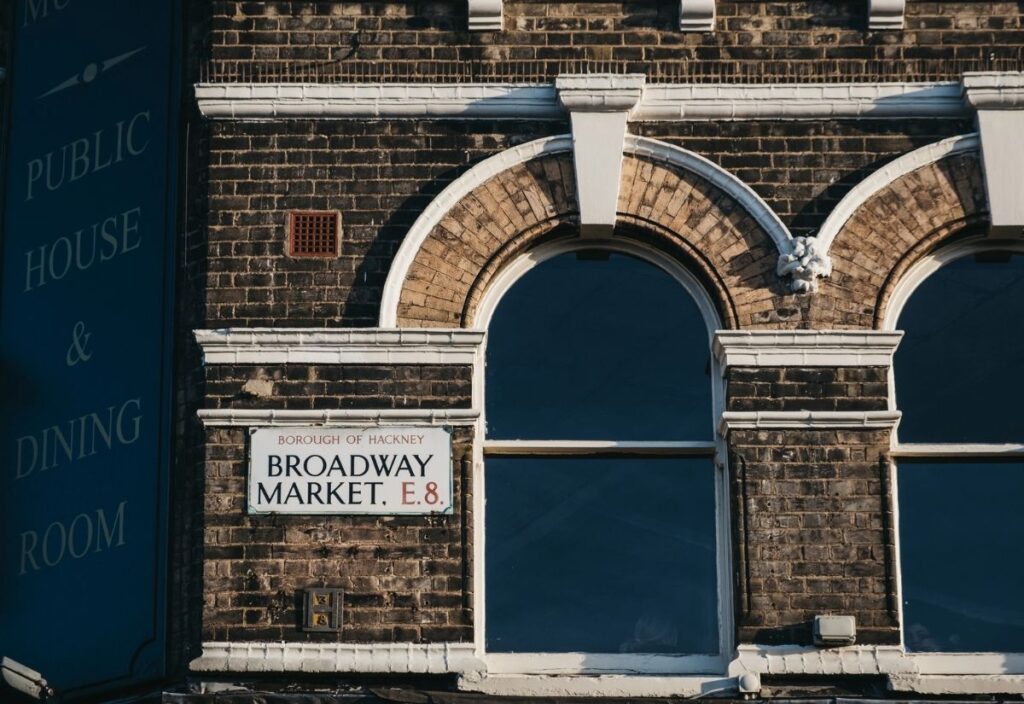 Brick building facade with arched windows, sign reads "Borough of Hackney, Broadway Market, E8." Partial text "Public House & Dining Room" visible.