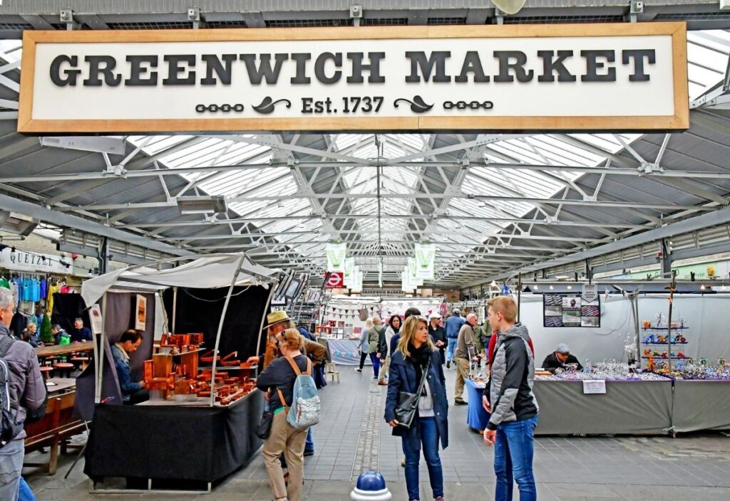 Bustling Greenwich Market interior with people browsing various artisan stalls under a large, hanging sign. The atmosphere is lively and vibrant.