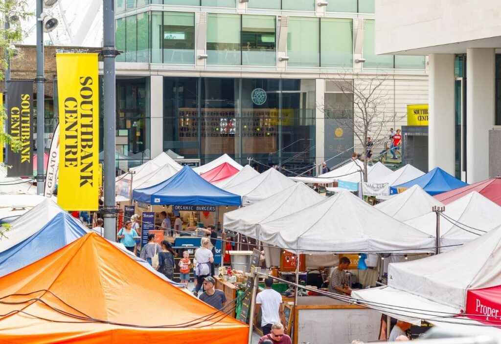 Outdoor market scene at Southbank Centre with colorful tents, bustling crowds, and a backdrop of a modern glass building. Energetic and vibrant atmosphere.