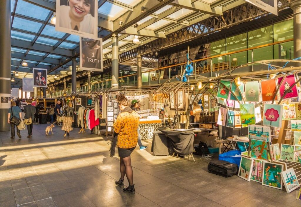 Indoor market scene at Spitalfields Market in London, showing various colorful stalls selling artwork, clothes, and crafts under a glass roof with people browsing and walking around.