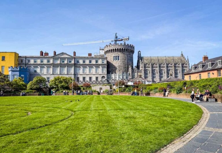 A sunny view of Dublin Castle, featuring a circular stone tower and historic buildings. Lush green lawn in the foreground, people sitting on benches.