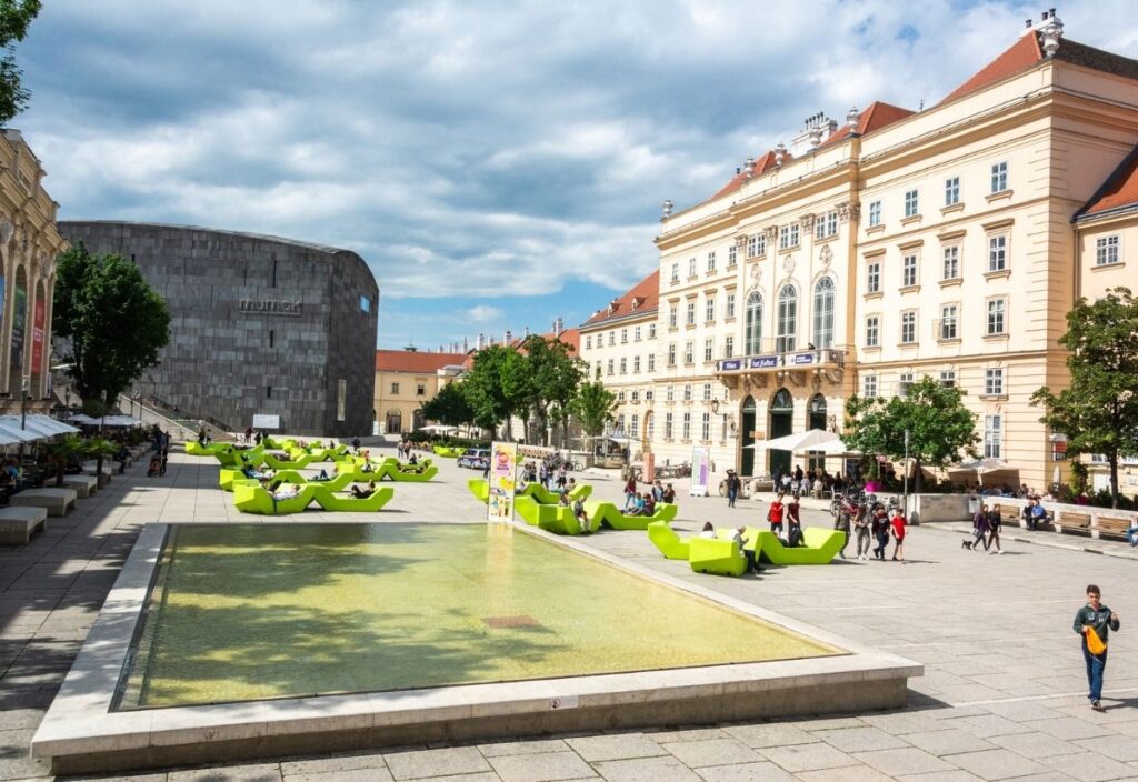 A bustling urban plaza with people walking and sitting on bright green lounge chairs. Historic buildings and a modern, grey structure frame the scene.