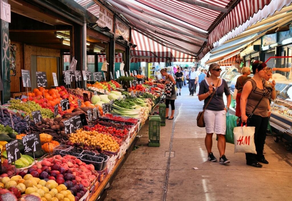 Bustling outdoor market with vibrant fruits and vegetables on display under striped canopies. Shoppers, casually dressed, browse and chat energetically.