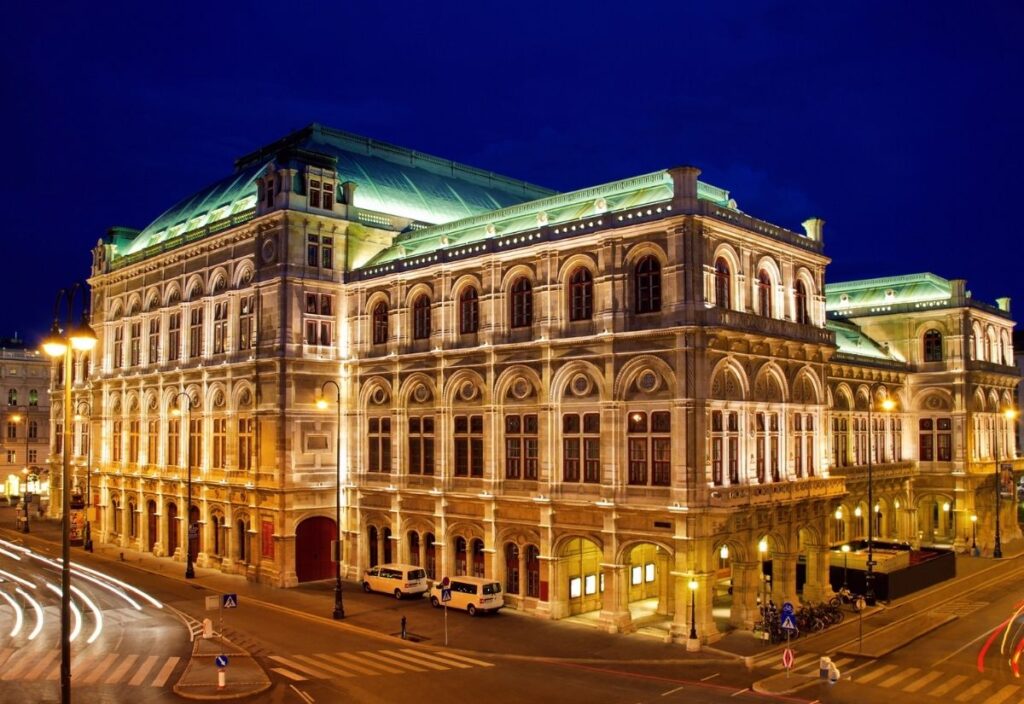 Illuminated opera house at night, showcasing ornate architecture with arched windows and a green roof. Nearby street lights and light trails from passing cars add life.
