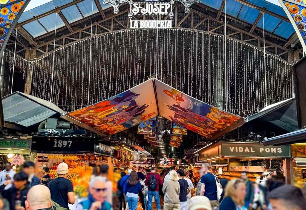 The image shows the lively entrance to La Boqueria Market in Barcelona, with its colorful stained glass and bustling crowd inside.