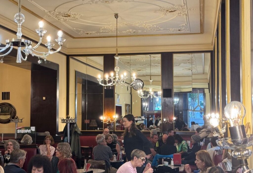 The image showcases a charming café with elegant chandeliers, vintage artwork on the wall, and a warm, inviting atmosphere. Guests are enjoying their time in a cozy, stylish setting.