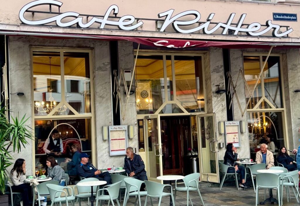 The image shows the exterior of Café Ritter, a charming café with outdoor seating. People are enjoying drinks at the tables, while a waiter can be seen inside. The café's name is prominently displayed above the entrance.