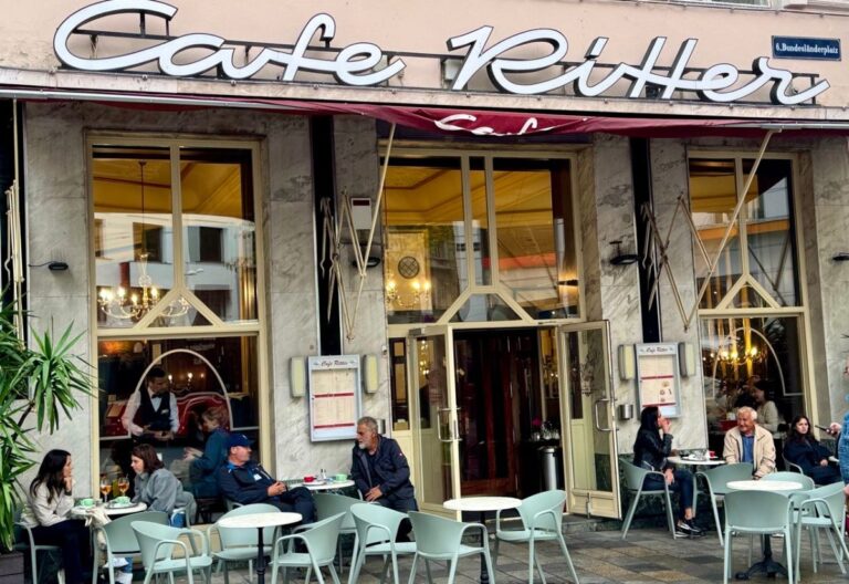 The image shows the exterior of Café Ritter, a charming café with outdoor seating. People are enjoying drinks at the tables, while a waiter can be seen inside. The café's name is prominently displayed above the entrance.
