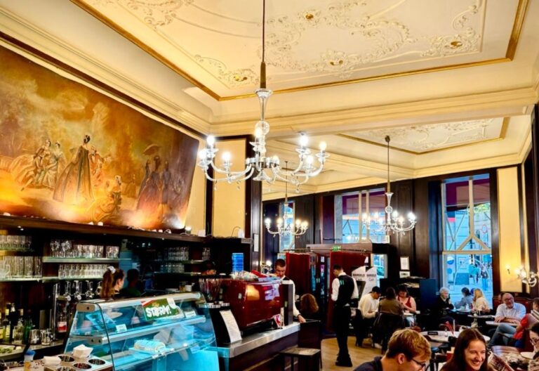 The image showcases a charming café with elegant chandeliers, vintage artwork on the wall, and a warm, inviting atmosphere. Guests are enjoying their time in a cozy, stylish setting.