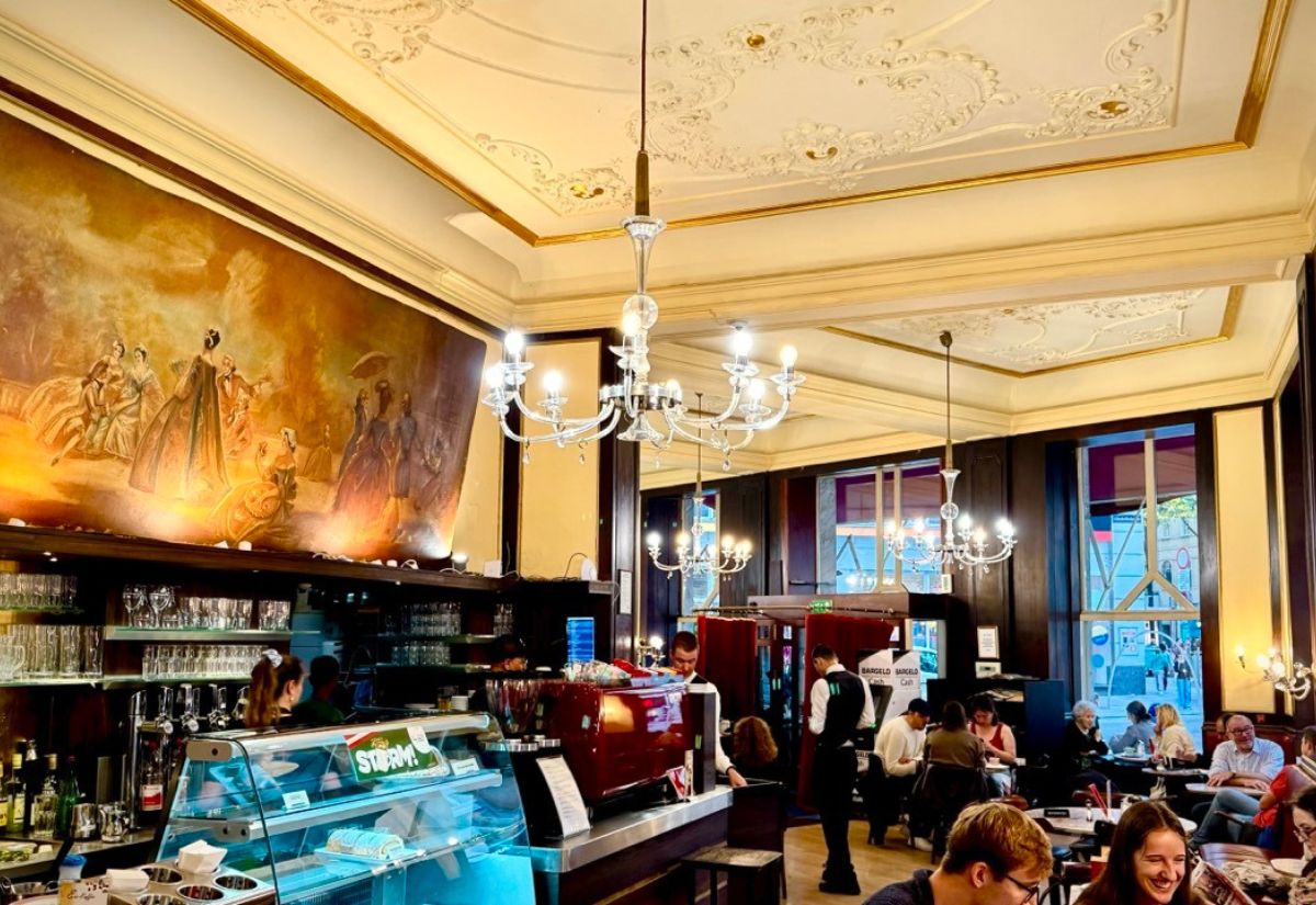 The image showcases a charming café with elegant chandeliers, vintage artwork on the wall, and a warm, inviting atmosphere. Guests are enjoying their time in a cozy, stylish setting.