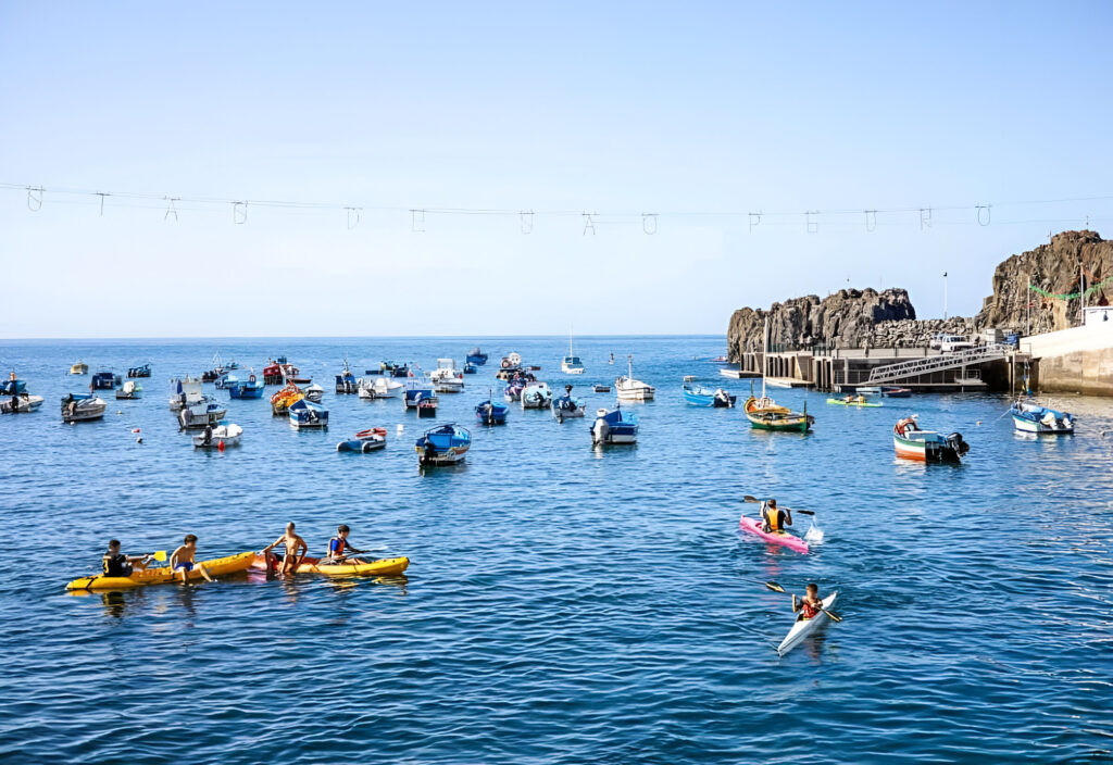 busy coastal harbor with colorful fishing boats anchored on calm blue water and people kayaking near the shore on a sunny day.