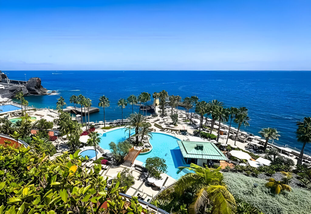 A scenic view of a coastal resort with palm trees, large swimming pools, sun loungers, and the blue ocean stretching to the horizon under a clear sky.