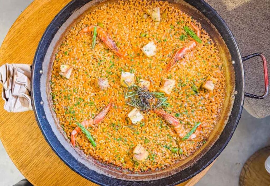 A large pan of seafood paella with shrimp, cubes of meat, and garnished with herbs and green onions on top of a rustic wooden table.