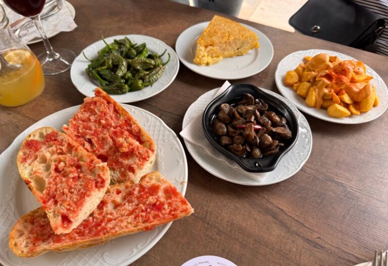 A table set with a variety of tapas including tomato-rubbed bread, grilled peppers, a slice of tortilla, marinated mushrooms, and fried potatoes with paprika, along with a glass of refreshing drink.
